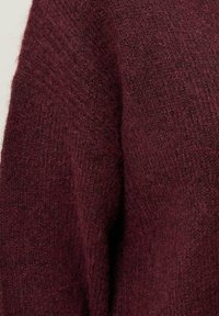 Primo piano di un maglione a maglia color bordeaux, caratterizzato da un tessuto morbido e testurizzato con un design ampio e rilassato e dettagli a costine lungo i bordi.