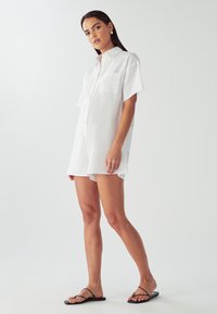 Bela obleka s kratkimi rokavi, ovratnikom, zapenjanjem na gumbe spredaj in prsnim žepom. Lahka tkanina z ohlapnim krojem. Črne sandale.