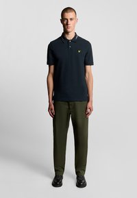 Polo shirt blu navy con colletto, dotato di un logo giallo sul petto. Abbinato a pantaloni verde oliva e stivali neri con lacci.