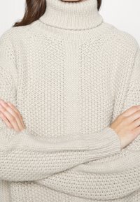 Jasnobeżowy dziergany sweter z golfem o teksturowanym wzorze, z prążkowanymi mankietami i luźnym krojem, prezentujący miękki, przytulny wygląd.