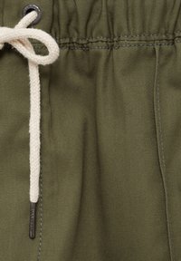 Gros plan sur un tissu vert olive avec une ceinture élastique et un cordon blanc tressé noué en un nœud avec des embouts métalliques et des œillets.