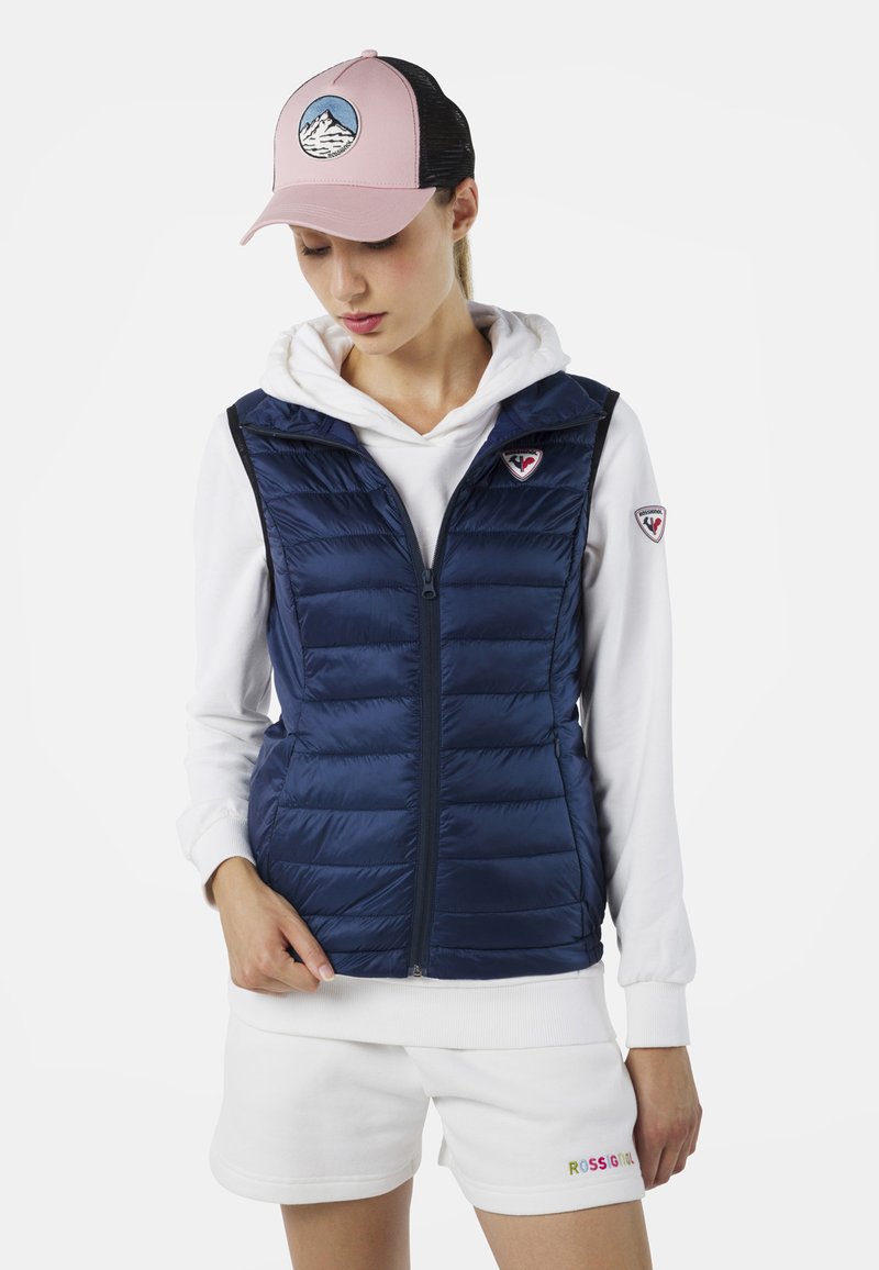 Marinblå puffer-väst med dragkedja och huva, lager på lager över en vit hoodie. Accentuerad med en liten logotyp på bröstet. Ljusrosa keps med bergdesign.