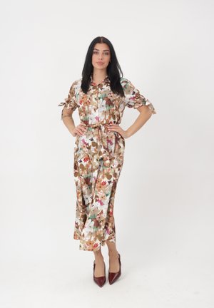 Frau steht mit Händen in den Hüften, trägt ein florales Midikleid mit gebundenen Ärmeln und bordeauxrote spitze Pumps vor weißem Hintergrund.