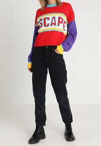 Czerwono-fioletowy sweter z napisem „ESCAPE”, ze ściągaczami w kolorze niebieskim i żółtym, zestawiony z czarnymi spodniami i czarnymi butami.
