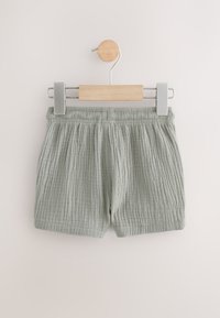 Pantaloncini di cotone verde chiaro con trama a costine, dotati di una vita elasticizzata e un design semplice, esposti su una gruccia di legno.