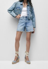 Ljusblå denimjacka med avslappnad passform, kombinerad med högmidjade shorts med fransar. Bruna espadrillos med ljusblå accenter.