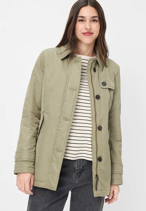 Frau trägt einen hellolivgrünen, geknöpften Mantel über einem weiß-schwarz gestreiften Hemd und dunkelgrauen Jeans, steht vor einem weißen Hintergrund.