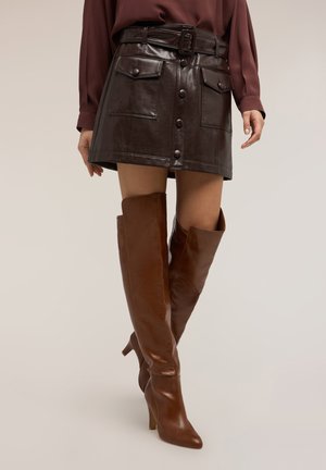 Donna che indossa una minigonna in pelle marrone scuro con tasche frontali e bottoni, abbinata a stivali alti in pelle marrone con tacco.