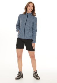 Blauwe fleece jas met rits, hoge kraag, twee zijzakken, gecombineerd met zwarte shorts en wandelschoenen. Soepele textuur en casual ontwerp.