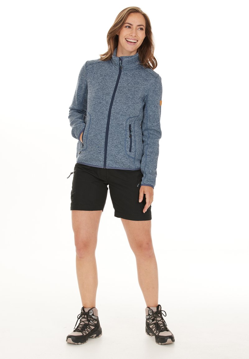 Blauwe fleece jas met rits, hoge kraag, twee zijzakken, gecombineerd met zwarte shorts en wandelschoenen. Soepele textuur en casual ontwerp.