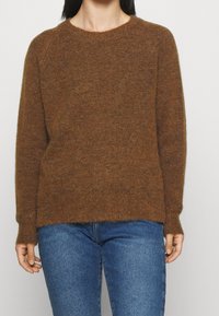 Suéter marrón de punto con cuello redondo y mangas largas. Textura suave, puños acanalados y corte relajado. Lleva puesto sobre unos vaqueros azules.