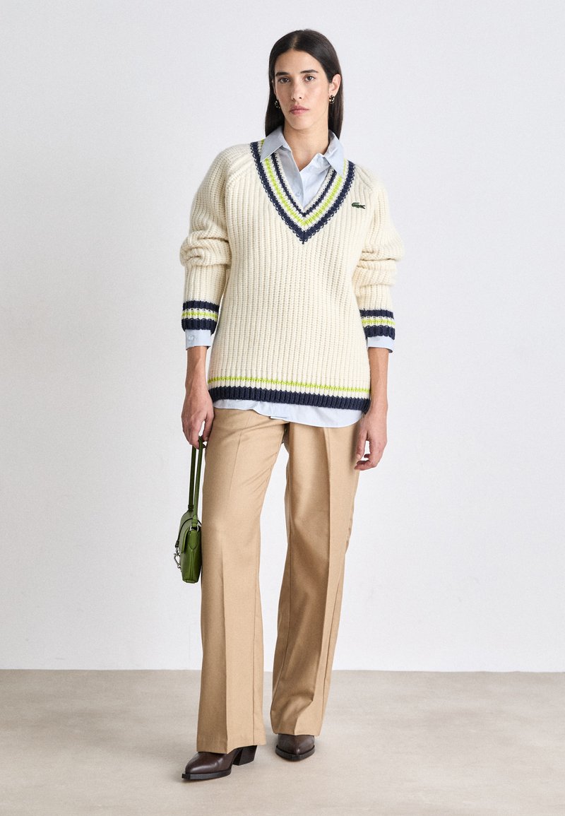 Pull crème surdimensionné avec col en V et rayures bleu marine/vert, associé à un pantalon beige. Le modèle tient un petit sac à main vert.