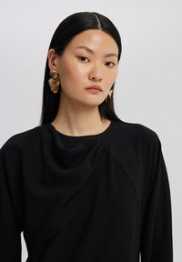 Svart topp med draperad halsringning och mjuk textur. Modellen bär gyllene blommörhängen. Bakgrunden är neutral grå.