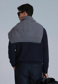 Grijze gebreide trui hangt over de schouders, met ribbelaccenten. Draagt het over een marineblauw shirt, met donkere broeken en een zwarte jas in de hand.
