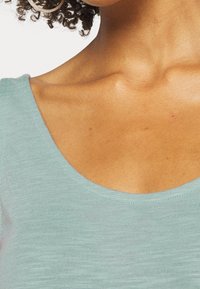 Ljus turkos kortärmad topp med rundad halsringning i ett mjukt, strukturerat material. Tyget har en avslappnad passform som framhäver nyckelbenområdet.