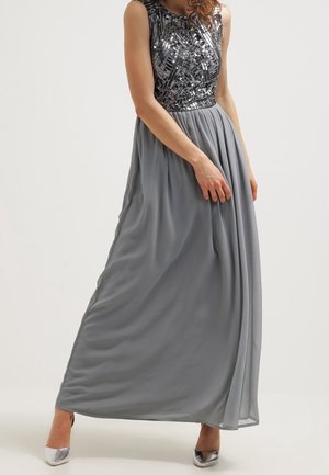 Kvinde iført en ærmeløs kjole med en sølvglitrende korset og et flydende gråt skørt, kombineret med sølvfarvede høje hæle.