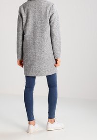 Abrigo gris texturizado con una silueta recta, combinado con jeans ajustados azules y zapatillas blancas; vista trasera muestra mangas largas y un cuello.