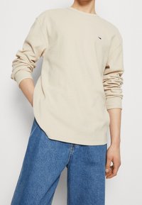 Beżowa koszula z długim rękawem, okrągłym dekoltem i prążkowaną strukturą. Posiada małe kolorowe logo na piersi. Połączona z luźnymi niebieskimi dżinsami.