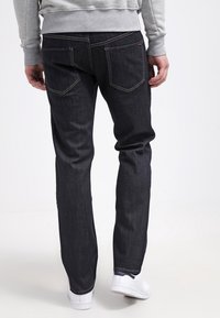 Jeans de denim oscuro con un ajuste recto, que presentan costuras visibles, dos bolsillos traseros, llevados con zapatillas blancas y un suéter gris.