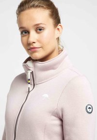 Giacca in pile rosa chiaro con zip e colletto alto, caratterizzata da un logo bianco e cordini regolabili. Tessuto liscio con rivestimento interno morbido.