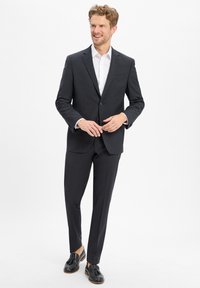 Completo grigio scuro con un design aderente, abbinato a una camicia bianca e mocassini neri. Tessuto morbido con una leggera trama. Aspetto sartoriale e curato.