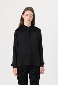 Zwarte langemouwen shirt met textuurplooien op de schouder, voorzien van een ronde kraag en een knoopsluiting aan de voorkant. Gecombineerd met gestreepte zwarte broek.