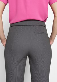 Personne portant un pantalon gris à carreaux ajusté avec une poche passepoilée à l’arrière et un haut rose vif à manches courtes, les mains posées sur les hanches.