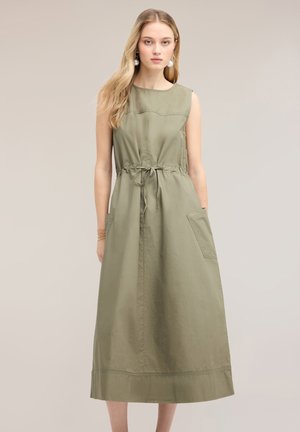 Femme aux longs cheveux blonds portant une robe midi sans manches vert olive avec poches avant et ceinture à la taille, debout devant un fond uni.