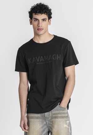 Jonge man met krullend haar, gekleed in een zwart Kavanagh T-shirt en lichtgewassen jeans, staand met één hand in de zak tegen een witte achtergrond.