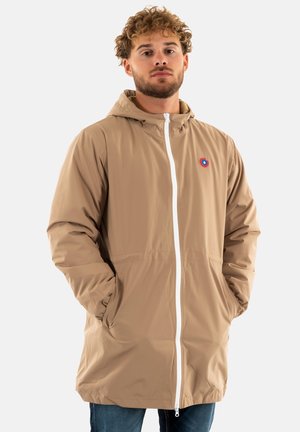 Veste imperméable beige avec capuche, fermeture éclair blanche et un petit logo rouge et bleu sur la poitrine. Comprend des poches latérales et des poignets élastiques.