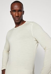 Chemise à manches longues de couleur crème en tissu léger, avec uncol arrondi et une texture subtile, ornée d'un détail logo sur la manche.