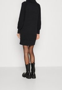Robe noire en maille avec un col haut, des détails côtelés sur les manches et une coupe décontractée, associée à des bottines noires brillantes.