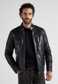 Marinblå läderjacka med dragkedja, dekorativ sömnad och sidofickor med dragkedja. Har en ståkrage och en slät, glänsande yta.