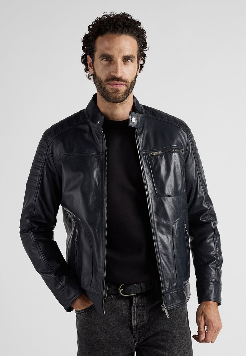 Marinblå läderjacka med dragkedja, dekorativ sömnad och sidofickor med dragkedja. Har en ståkrage och en slät, glänsande yta.