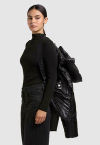 Jonge vrouw in een zwarte coltrui en jeans draagt een zwart jasje met het "KHUJO ORIGINAL" logo over haar schouder.