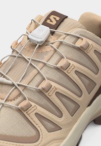 Gros plan d'une chaussure de sport beige et marron avec tissu en maille, superpositions synthétiques, et système de laçage à tirage rapide avec verrou en plastique.