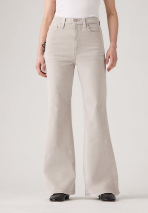 Personne portant un pantalon beige clair taille haute à jambes larges avec des poches avant et des chaussures noires pointues, debout devant un fond uni.