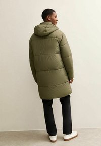 Olivengroene donsjas met capuchon, voorzien van horizontale doorgestikte secties. Draagt over een zwarte broek en witte schoenen, gezien van achteren.