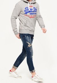 Szara bluza z kapturem z czerwono-niebieskim nadrukiem, przetarte niebieskie dżinsy i białe trampki z czerwonymi akcentami. Styl casual, dopasowany fason.