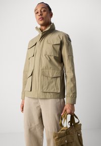 Jachetă utilitară khaki cu multiple buzunare frontale și buzunar cu fermoar pe manșetă. Purtați peste pantaloni bej deschis, ținând o geantă de umăr dintr-un material asortat.