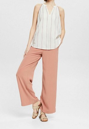 Kobieta nosząca białą, bezrękawnikową bluzkę w pionowe paski, szerokie różowawe spodnie i beżowe sandały, stojąca na jednolitym tle.