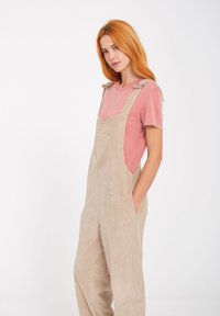 Beige corduroy overalls med brede ben, med justerbare stropper og en frontlomme, båret over en lyserød t-shirt med korte ærmer.