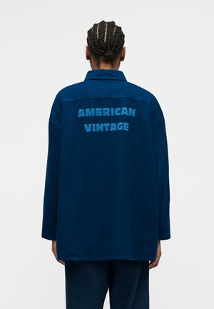 Persoană cu părul împletit purtând o jachetă albastru închis cu inscripția „AMERICAN VINTAGE” pe spate, stând în fața unui fundal simplu.