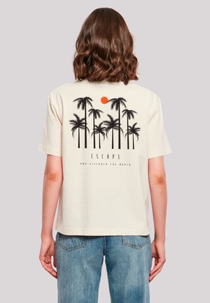Person with shoulder-length brown hair stands facing away, wearing cream t-shirt with black palm trees and orange sun graphic and blue jeans.