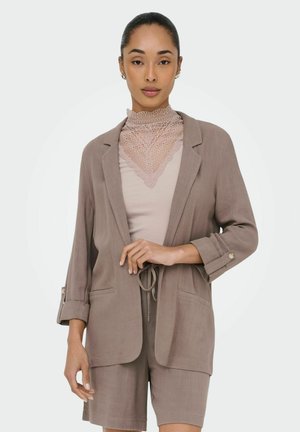 Kvinde med trukket hår tilbage iført taupe blazer og matchende shorts over en beige blondehøjhalset top, poserende med den ene hånd nær taljen.