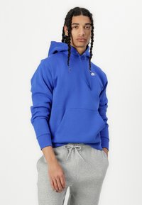 Casaco de polar azul com um bolso frontal, cordão ajustável e logotipo branco no peito. Combinado com calças de moletom cinza claro.