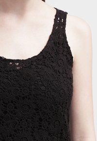 Top sin mangas de encaje negro con un patrón floral, escote redondo y tirantes anchos. La textura es intrincada y tiene un aspecto transparente.