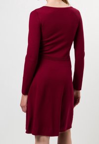 Vestido burdeos de manga larga con corsé ajustado y falda acampanada, que presenta un detalle de cinturilla texturizada y un acabado de tela suave.