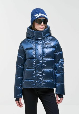 Femme portant une doudoune bleue brillante avec capuche, un bonnet bleu assorti, de grandes lunettes de soleil noires et un pantalon noir sur un fond uni.