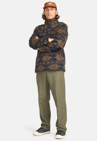 Pull en polaire avec un motif géométrique marron, bleu marine et moutarde ; col montant ; associé à un pantalon vert olive et des baskets noires.